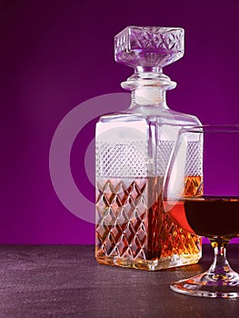glass textured square bottle and glass goblet with amber alcohol brandy scotch whiskey. violet background