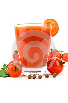 A glass of tomato juice with a tomato slice on the edge. Tomatoes, parsley and allspice are spread around