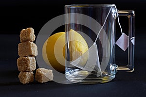 A glass transparent mug with a pyramid tea bag, fresh yellow lemon and a tower of brown cane unrefined sugar cubes. Dark