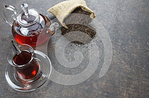 Glass Turkish brewed black tea and glass teapot with dry black tea in burlap sack on black rustic table