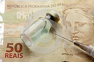 Glass vaccine bottle with liquid and syringe needle over a brazilian 50 Reais bank note from close