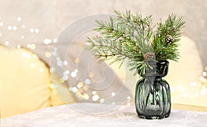 Glass vase with fir branches on table with tablecloth in living room with sofa and Christmas lights