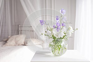 Glass vase with lilac and white floweers  in light cozy bedroom interior. White wall, bed with white linen, light blanket or plaid