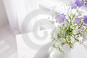 Glass vase with lilac and white floweers  in light cozy bedroom interior. White wall, sunlight from window, copy space