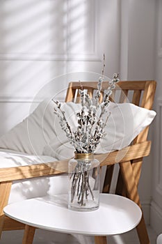 Glass vase with pussy willow tree branches on table near armchair