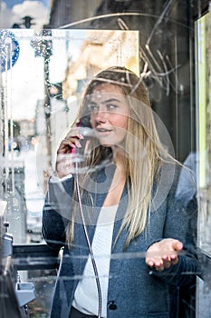 Through glass view of young woman talking on public telephone in payphone box