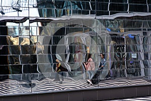 In the glass wall of a modern shopping center, broken reflections of people