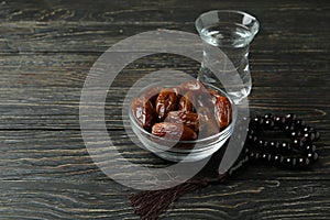 Glass of water, bowl of dates and rosary on wooden background