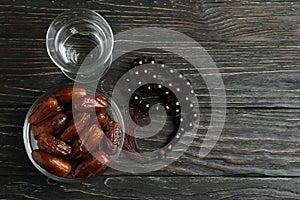 Glass of water, bowl of dates and rosary on wooden background