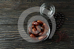 Glass of water, bowl of dates and rosary on wooden background