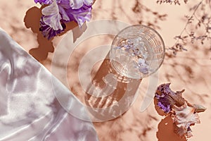 A glass with water and iris flowers on pastel background with shadows and white silk cloth. Summer refreshment concept. Sunlit