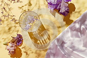 A glass with water and iris flowers on pastel yellow background with shadows and white silk cloth. Summer refreshment concept.