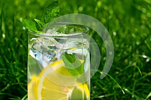 A glass of water with lemon and mint on the background of fresh summer green grass. Cooling drink.
