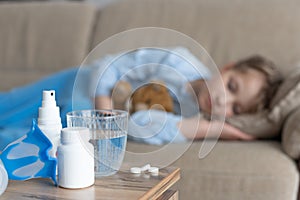 A glass of water, a medication and a mask for inhalation on the table and in the background out of focus a sick girl on the couch