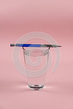 A glass of water and a pencil on a pink background. A full glass of drinking water. Blue and silver metallic pencil. Kurp plan.