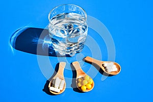 Glass of water and pills on wooden spoon on blue background with hard light and shadows. Creative. Vitamins and prebiotics, photo
