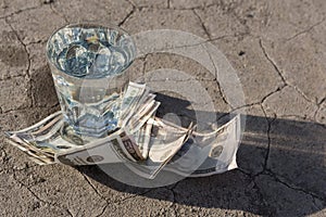 Glass with water stands on paper dollar bills and on dry ground with cracks, concept