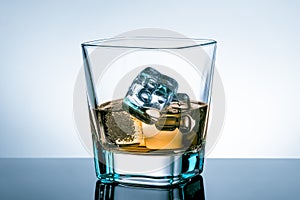 Glass of whiskey with ice cubes on bar table with reflection on light blue tint background