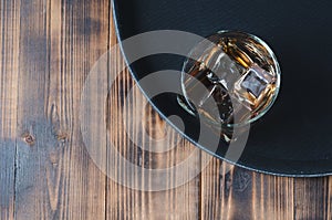 Glass of whiskey with ice cubes on a black tray and wooden table/Glass of whiskey with ice cubes on a black tray and wooden table