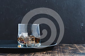 Glass of whiskey with ice cubes on a black tray and wooden table/Glass of whiskey with ice cubes on a black tray and wooden table