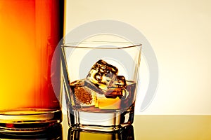 Glass of whiskey with ice cubes near bottle on table with reflection, warm tint atmosphere