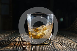 Glass of whiskey with ice cubes on a wooden table. An old tabletop with light and a glass of strong drink.