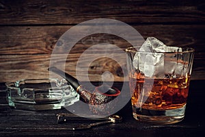 Glass of whiskey with ice and pipe on a wooden background.