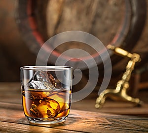 Glass of whiskey on top of wooden barrel and wood barrels in cellar at the background. Sun ray falling down on it surface