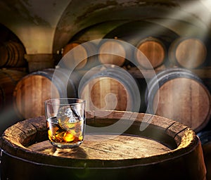 Glass of whiskey on top of wooden barrel and wood barrels in cellar at the background. Sun ray falling down on it surface