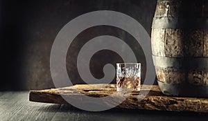 Glass of whisky or bourbon in ornamental glass next to a vinatge wooden barrel on a rustic wood and dark background