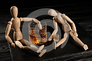 Glass of whisky and two mannequins on wooden table against black background. Alcoholism concept