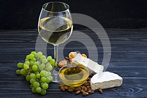 A glass of white wine with cheese cuts, figs, nuts, honey, grapes on a dark rustic wooden boards background