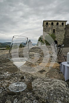 Glass of white wine in restaurant across ancient castle wall, restaurant terrace and mountains view. Langhirano, Italy. Travel