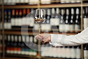 Glass of wine filled with white wine in female hand on cellar and bottles shelf background, close up with a free copy photo