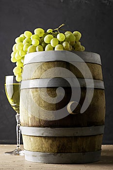 a glass of wine an oak barrel and a bunch of white grapes on a dark background