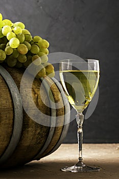 a glass of wine an oak barrel and a bunch of white grapes on a dark background