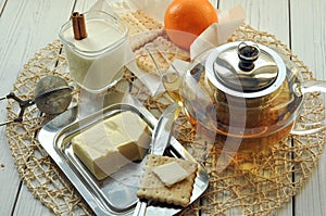 A glass of yogurt, butter, a pot of tea, pastry and fresh fruits on a light surface