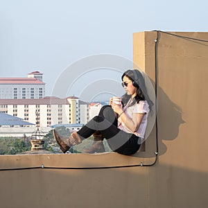 Glasses Asian woman is listening music via headset, drinking coffee on rooftop barricade cement not dangerous because the