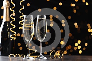 Glasses and bottle of champagne with serpentine streamers against blurred lights on black background. Space for text