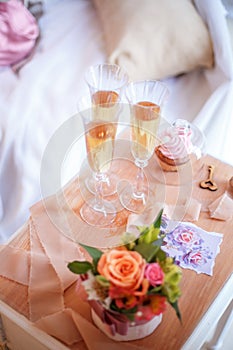 Glasses with champagne, cake, flowers, wooden key and tapes on the bedside table