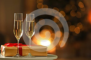 Glasses with champagne and gift on table in room, bokeh effect. Space for text