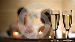 Glasses of champagne on foreground, lovers having pleasure pastime in bathroom