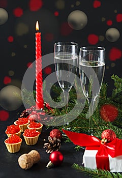 Glasses of champagne, red candle, tartlets with red caviar, gift box with red ribbon , fir branches  on a black background bokeh
