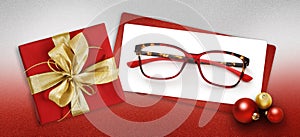 Glasses christmas gift card, red box with shiny golden ribbon bow, white ticket and eyewear near xmas balls on glittering red