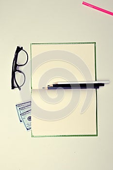Glasses on a clean notebook pages on white table background pink red blue black white color pencils dollars money is all