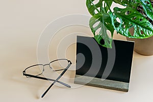 Glasses, monstera plant and black blank on the workdesk. Look above at an angle. Mock up photo, offer to display your text or sign