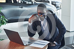 In glasses. Office worker is sitting in autosalon by table with laptop