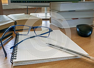 Glasses and a pen on a blank sheet of notebook. Books, a monitor, and other items for work are on the table.