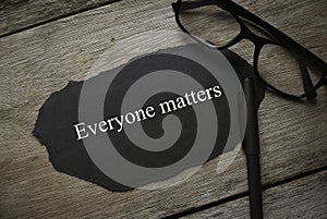 Glasses,pen and piece of black paper written with Everyone Matters on wooden background