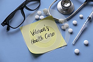 A glasses, pills, syringe, stethoscope and yellow memo sticky note written with Veteran's Health Care on blue background